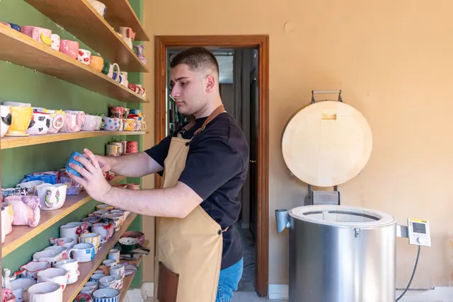 Young male potter organizing colorful handmade ceramic mugs in a pottery studio with a kiln. Projects that teach one kiln skill at a time