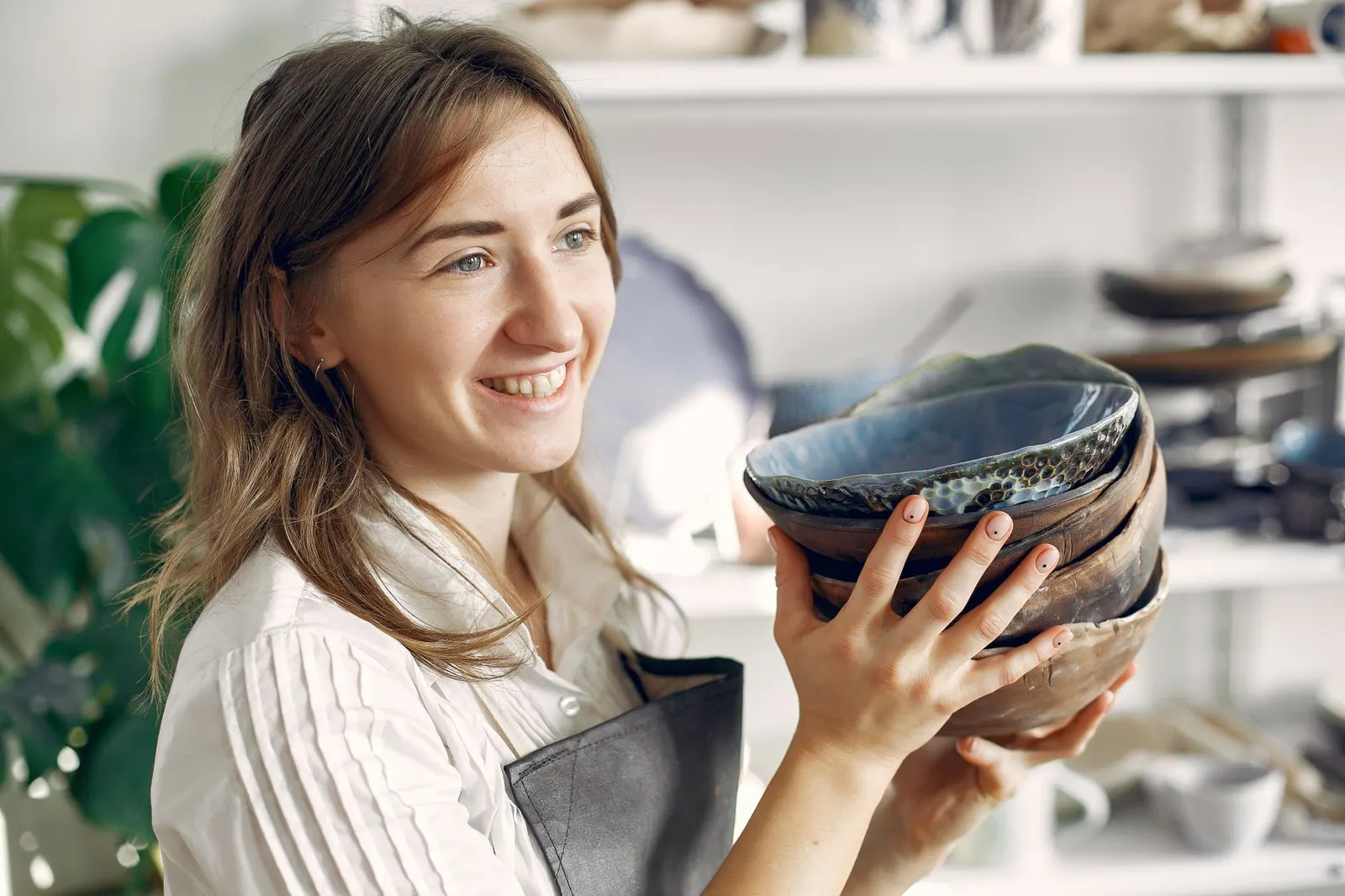 Cheerful young female ceramist with handmade ceramic bowls standing in light sunny modern pottery and looking away. What to buy first to avoid the usual studio mistakes